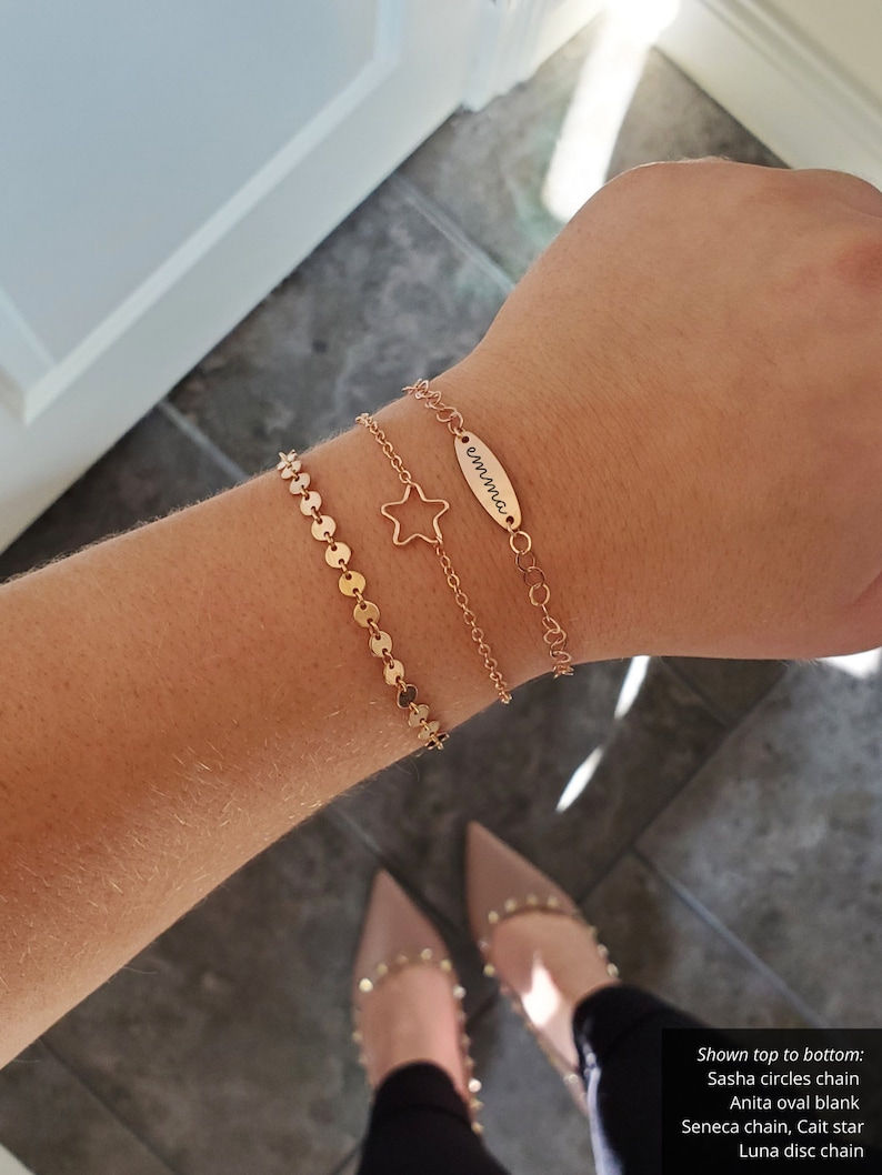 Puede incluir: Tres pulseras de oro apiladas en una mu&ntilde;eca. La pulsera superior es una cadena fina con una peque&ntilde;a etiqueta ovalada que dice "emma". La pulsera del medio es una cadena delicada con un dije de estrella. La pulsera inferior es una cadena de peque&ntilde;os c&iacute;rculos dorados.