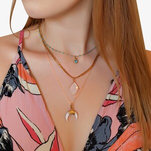 May include: A layered necklace set featuring gold chains and pendants. The top necklace has small turquoise beads and a blue charm. The middle necklace has a diamond-shaped pendant. The bottom necklace has a white horn pendant. The text in the image says: Nellie necklace, Elise necklace, Sloane necklace.