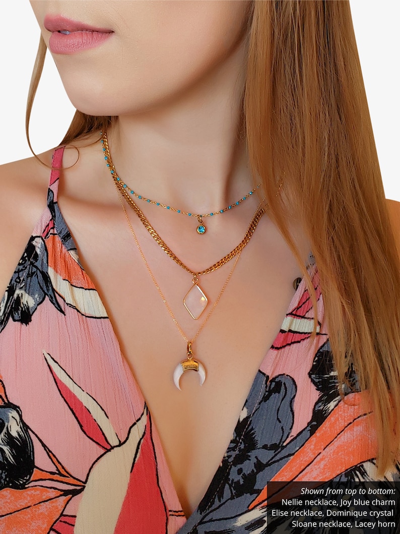 May include: A layered necklace set featuring gold chains and pendants. The top necklace has small turquoise beads and a blue charm. The middle necklace has a diamond-shaped pendant. The bottom necklace has a white horn pendant. The text in the image says: Nellie necklace, Elise necklace, Sloane necklace.