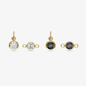 May include: Three gold charms with a round bezel setting. The charms feature a clear, white stone, a clear, white stone with a connector loop, and a black stone with a connector loop.