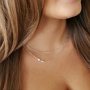 May include: Two gold necklaces layered on a person's neck. The top necklace is a delicate chain with a small opal pendant. The bottom necklace is a thicker chain with a paperclip design.