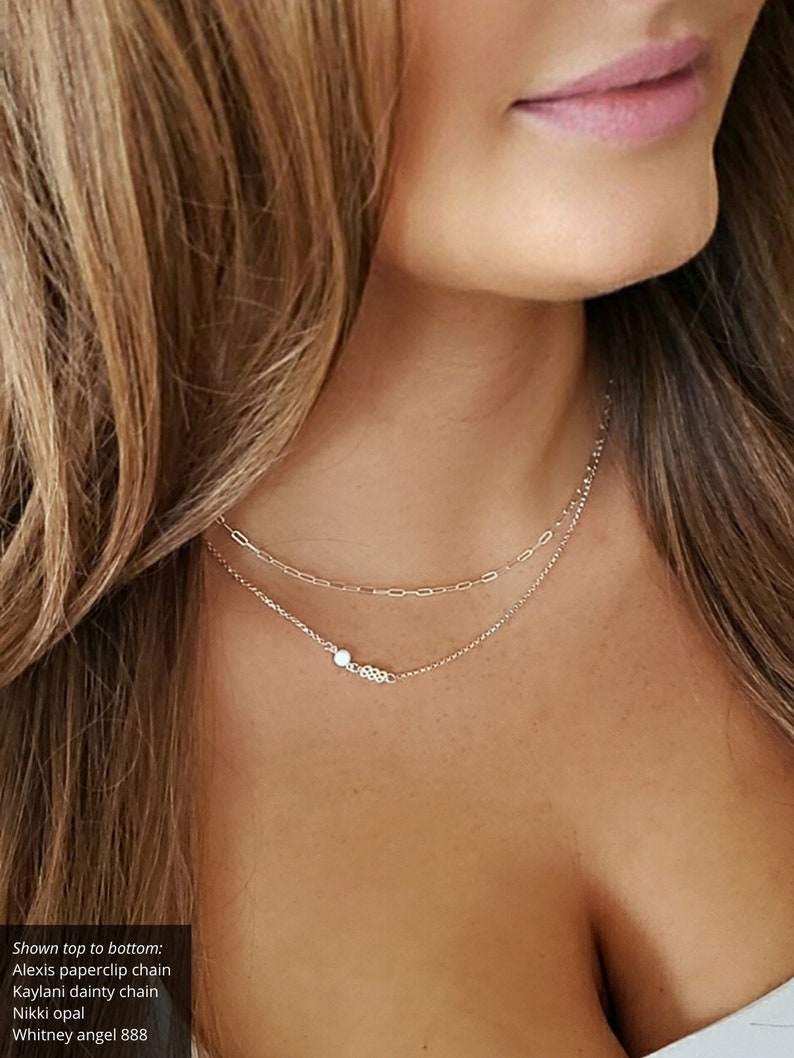 May include: Two gold necklaces layered on a person's neck. The top necklace is a delicate chain with a small opal pendant. The bottom necklace is a thicker chain with a paperclip design.