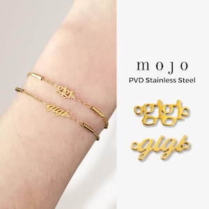 May include: Two gold-colored bracelets with the name "gigi" in cursive script. The bracelets are on a wrist. The image also shows the word "mojo" and "PVD Stainless Steel" in black text.