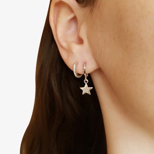 May include: A silver star charm dangling from a silver hoop earring. A smaller silver hoop earring with clear stones is worn higher on the ear.
