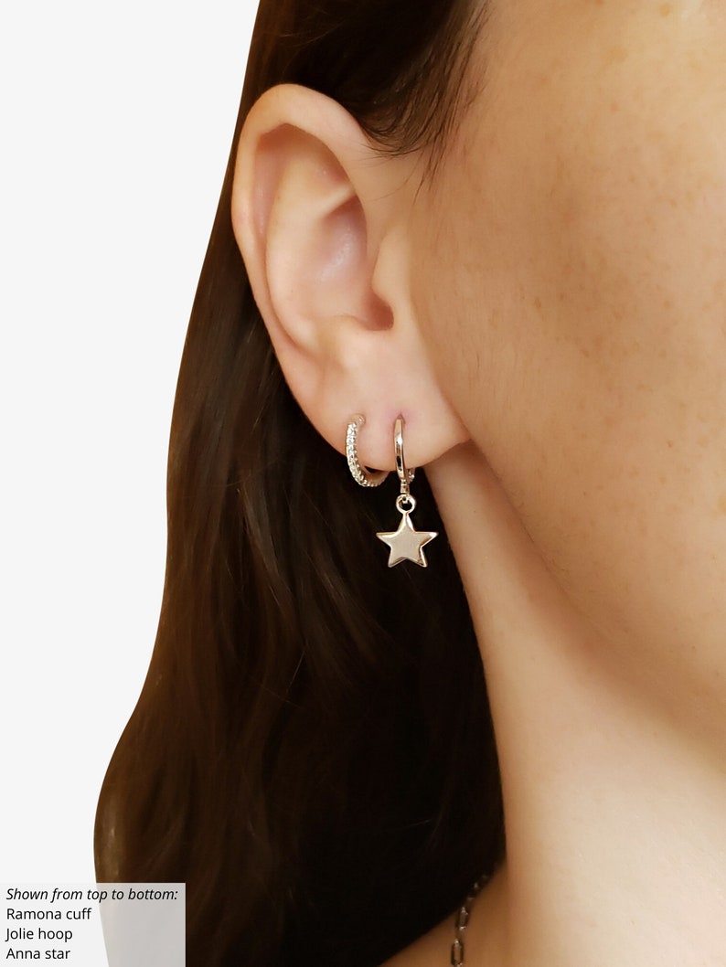 May include: A silver star charm dangling from a silver hoop earring. A smaller silver hoop earring with clear stones is worn higher on the ear.