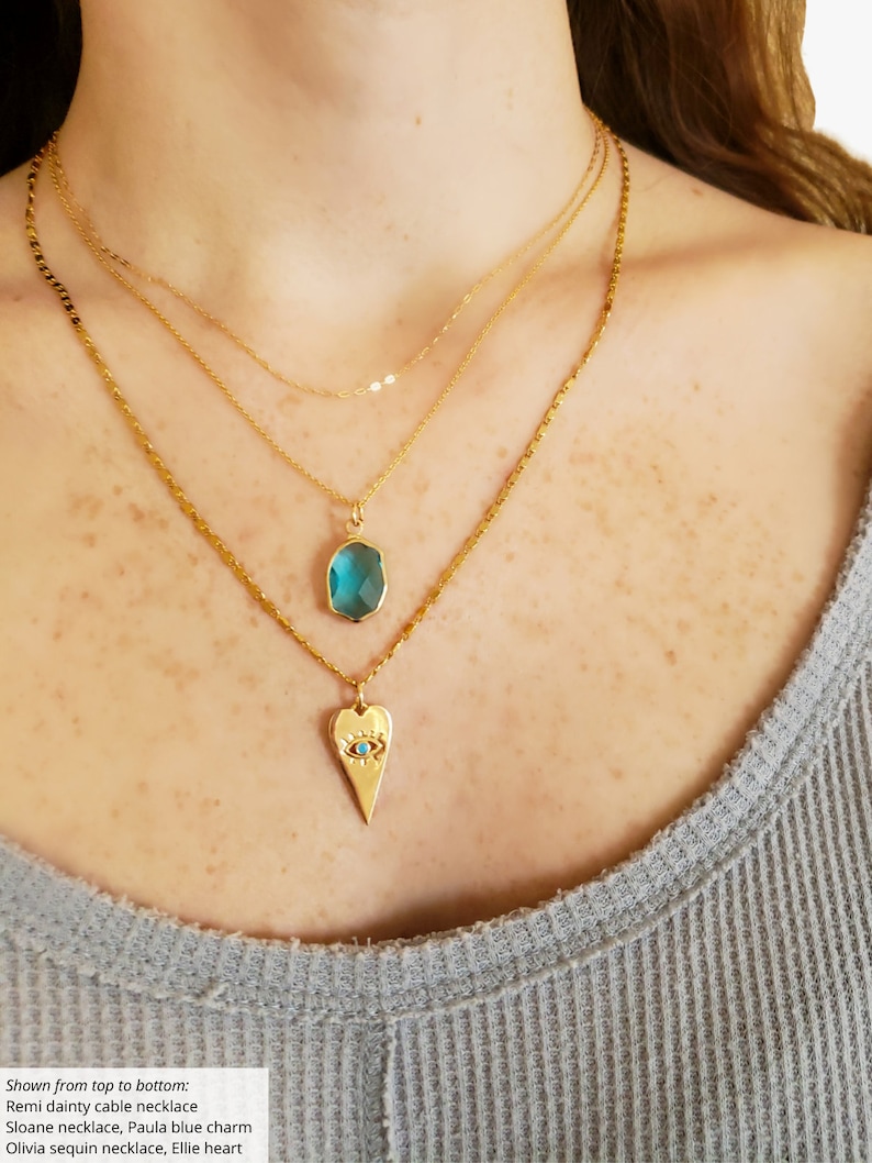 May include: Three gold necklaces layered on a person's neck. The top necklace is a delicate chain with a small oval-shaped blue gemstone pendant. The middle necklace is a thicker chain with a heart-shaped pendant featuring a blue eye design. The bottom necklace is a chain with a sparkly, sequin-like texture.