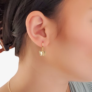 May include: Gold butterfly dangle earrings with a hoop design. The earrings are shown on a model's ear.