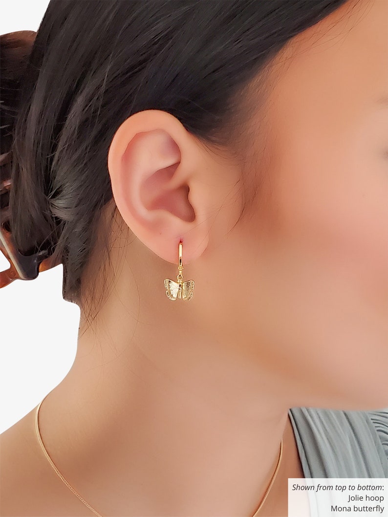 May include: Gold butterfly dangle earrings with a hoop design. The earrings are shown on a model's ear.