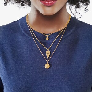 May include: A woman wearing a blue shirt with five gold necklaces. The necklaces feature a coin, a heart, a locket, and two small charms.