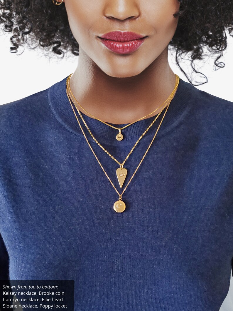 May include: A woman wearing a blue shirt with five gold necklaces. The necklaces feature a coin, a heart, a locket, and two small charms.