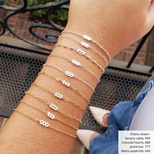 May include: A person's wrist wearing a stack of gold and silver chain bracelets with numbers on them. The numbers on the bracelets are 999, 888, 777, 666, 555, 444, 333, 222, 111, and 000. The bracelets are all different styles and lengths. Chains shown: Seneca cable, 999, Cressida hearts, 888, Jackie bar, 777, Alexis paperclip, 666, Kaylani dainty, 555, Melanie sequin, 444, Alexis paperclip, 333, Cressida hearts, 222, Kaylani dainty, 111, Seneca cable, 000.
