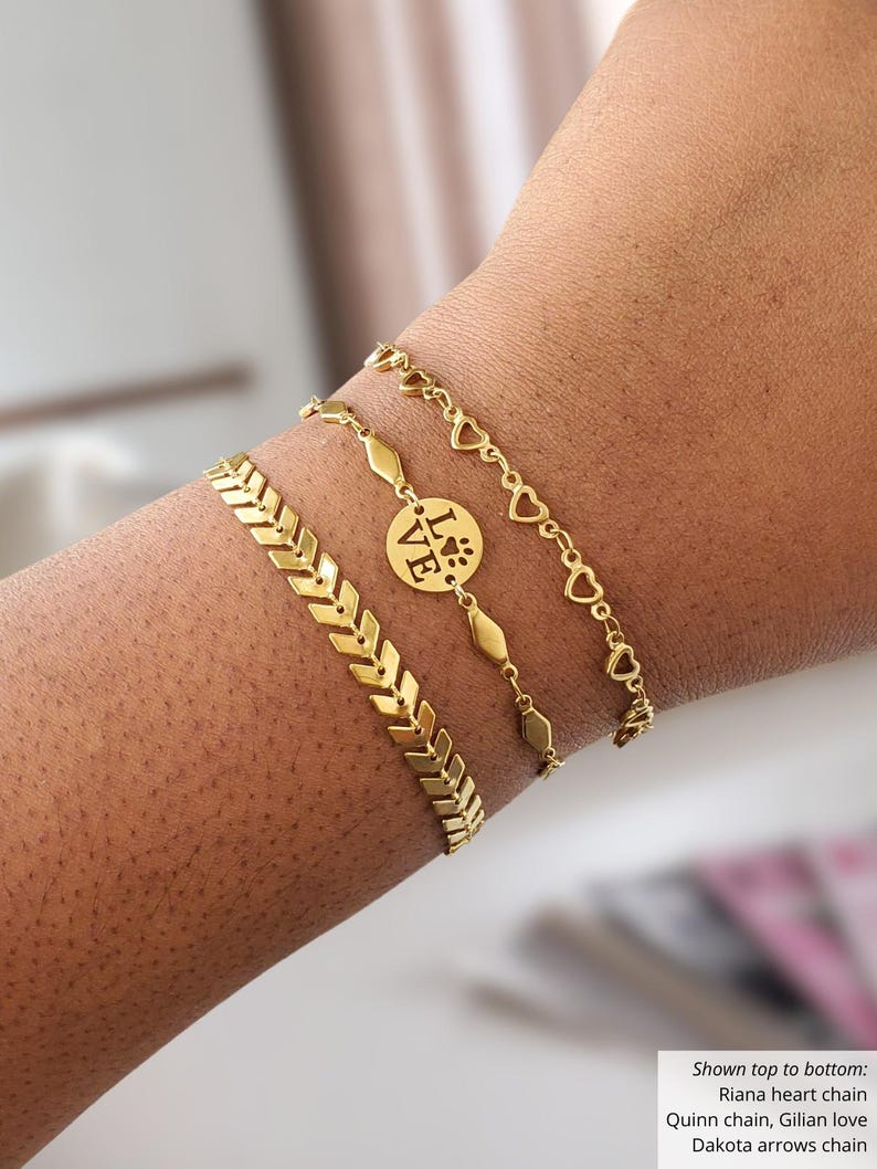 May include: Gold-colored bracelets stacked on a wrist. The bracelets include a heart chain, a chain with a circular charm that says "LOVE" and a paw print, a chain with geometric shapes, and an arrow design. Jewelry.