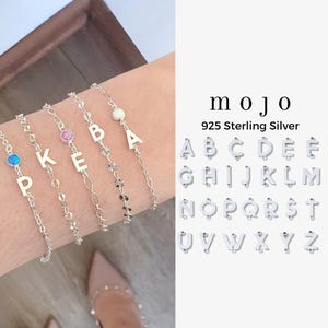 May include: Silver charm bracelets with the letters P, K, E, B, and A. The bracelets are on a wrist, with a white background. The word "mojo" and "925 Sterling Silver" are in the image, along with the alphabet.