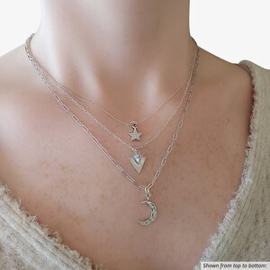 May include: A layered necklace set featuring three silver-toned chains. The top chain has a star pendant, the middle chain has a triangular pendant, and the bottom chain has a crescent moon pendant. Text reads: Shown from top to bottom: Sage necklace Anna star Cali necklace Orla triangle Vera necklace Melissa moon.