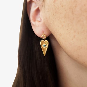 May include: Gold heart-shaped earring with a blue evil eye charm. The earring is shown on a model's ear.