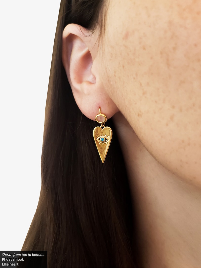May include: Gold heart-shaped earring with a blue evil eye charm. The earring is shown on a model's ear.