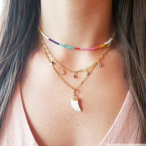 May include: A layered necklace set featuring a beaded choker in rainbow colors, a gold chain necklace with gemstone charms, and a pendant with a white horn design. The necklaces are displayed on a person wearing a pink top.