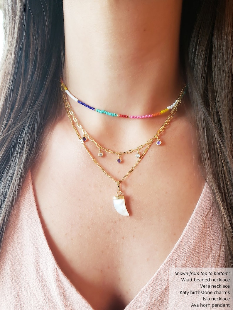 May include: A layered necklace set featuring a beaded choker in rainbow colors, a gold chain necklace with gemstone charms, and a pendant with a white horn design. The necklaces are displayed on a person wearing a pink top.