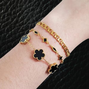 K&ouml;nnte beinhalten: Drei goldene Armb&auml;nder werden gezeigt. Das obere Armband ist eine dicke Gliederkette. Das mittlere Armband hat rautenf&ouml;rmige Glieder. Das untere Armband hat schwarze Kleeblatt-Anh&auml;nger. Der Text im Bild lautet: Chelsea thick link chain, Tinsley clover diamond chain, Tiff black flower connectors.