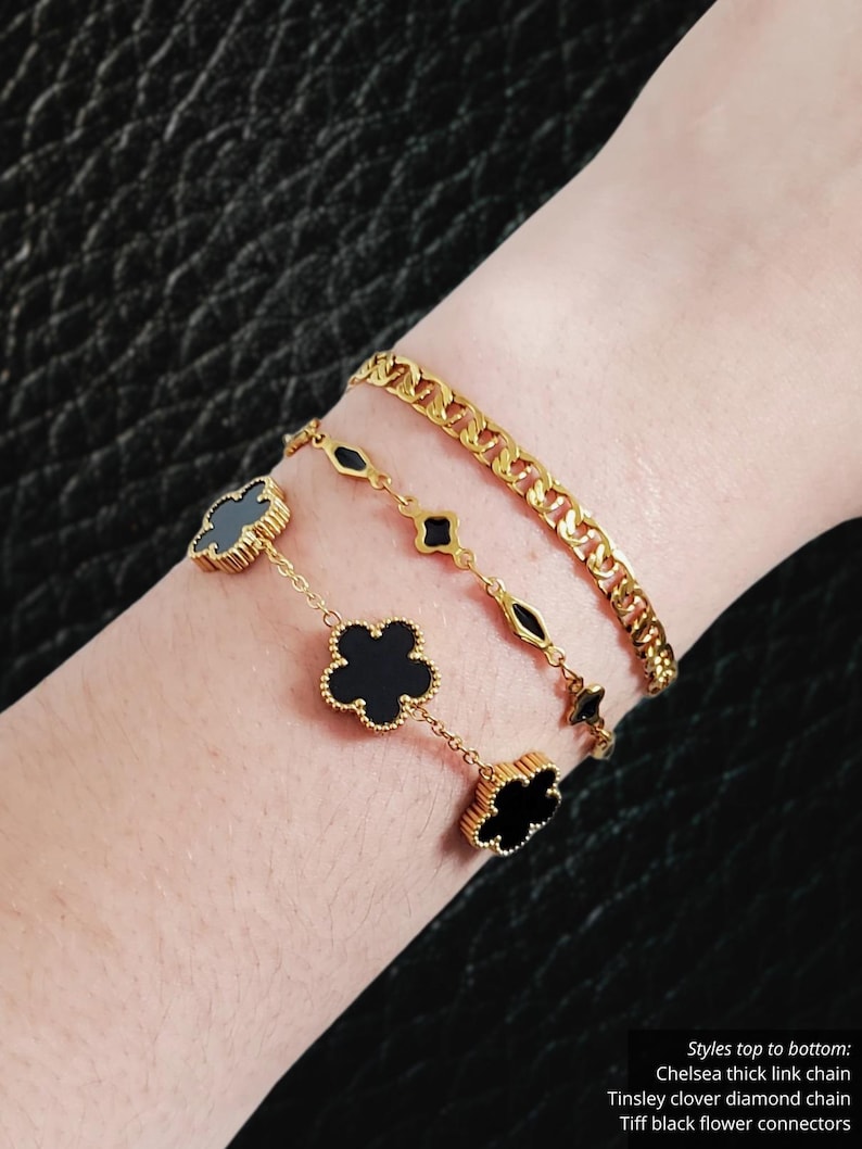 K&ouml;nnte beinhalten: Drei goldene Armb&auml;nder werden gezeigt. Das obere Armband ist eine dicke Gliederkette. Das mittlere Armband hat rautenf&ouml;rmige Glieder. Das untere Armband hat schwarze Kleeblatt-Anh&auml;nger. Der Text im Bild lautet: Chelsea thick link chain, Tinsley clover diamond chain, Tiff black flower connectors.