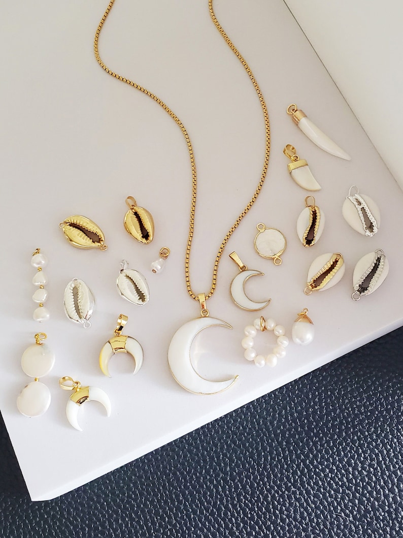 May include: A collection of gold-toned and white shell pendants, necklaces, and charms. The jewelry includes a gold chain necklace with a large white crescent moon pendant, and a pearl ring.