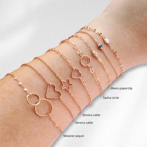 Puede incluir: Primer plano de la mu&ntilde;eca de una mujer con diez pulseras de oro con diferentes estilos de cadena y dijes, incluyendo un coraz&oacute;n, una estrella y un c&iacute;rculo. Las pulseras est&aacute;n etiquetadas con nombres como "Sasha circle", "Seneca cable" y "Melanie sequin".