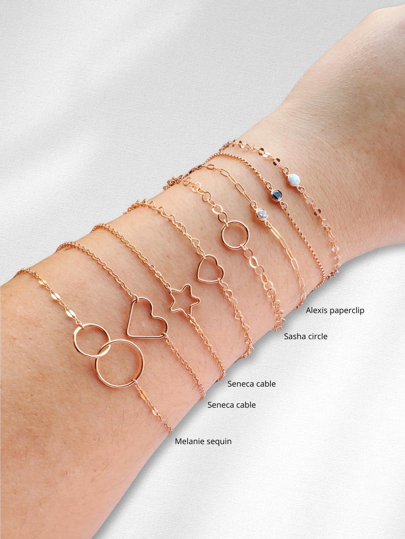 Puede incluir: Primer plano de la mu&ntilde;eca de una mujer con diez pulseras de oro con diferentes estilos de cadena y dijes, incluyendo un coraz&oacute;n, una estrella y un c&iacute;rculo. Las pulseras est&aacute;n etiquetadas con nombres como "Sasha circle", "Seneca cable" y "Melanie sequin".