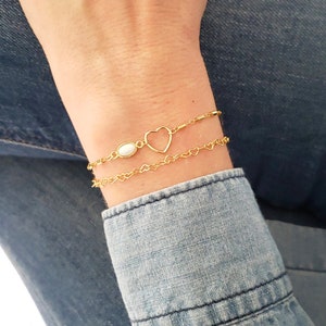 May include: A gold heart charm bracelet with a white opal connector. The bracelet is layered with two chains, one with a heart design and the other with a bar design.