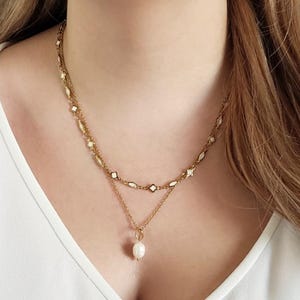 K&ouml;nnte beinhalten: Eine goldfarbene Halskette mit mehreren Schichten und einem Perlenanh&auml;nger. Die obere Schicht ist eine Kleeblattkette, die mittlere Schicht eine Kabelkette und die untere Schicht hat einen einzelnen Perlenanh&auml;nger. Die Halskette wird &uuml;ber einem wei&szlig;en Oberteil getragen.