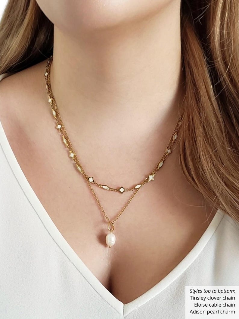 K&ouml;nnte beinhalten: Eine goldfarbene Halskette mit mehreren Schichten und einem Perlenanh&auml;nger. Die obere Schicht ist eine Kleeblattkette, die mittlere Schicht eine Kabelkette und die untere Schicht hat einen einzelnen Perlenanh&auml;nger. Die Halskette wird &uuml;ber einem wei&szlig;en Oberteil getragen.