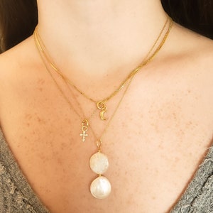 May include: A layered gold necklace set. The top necklace has a small crescent moon charm. The middle necklace has a small cross charm. The bottom necklace features two large, round, white pearl pendants. The text in the image reads: "Kelsey necklace, Elaina moon, Sloane necklace, Elaina cross, Sloane necklace, Rivka pearl."