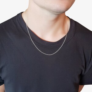 May include: A thin, silver-colored cable chain necklace with small, uniform oval links. The necklace rests against the neck, featuring a simple, delicate design with a polished metallic finish that catches the light, worn over a plain black t-shirt.
