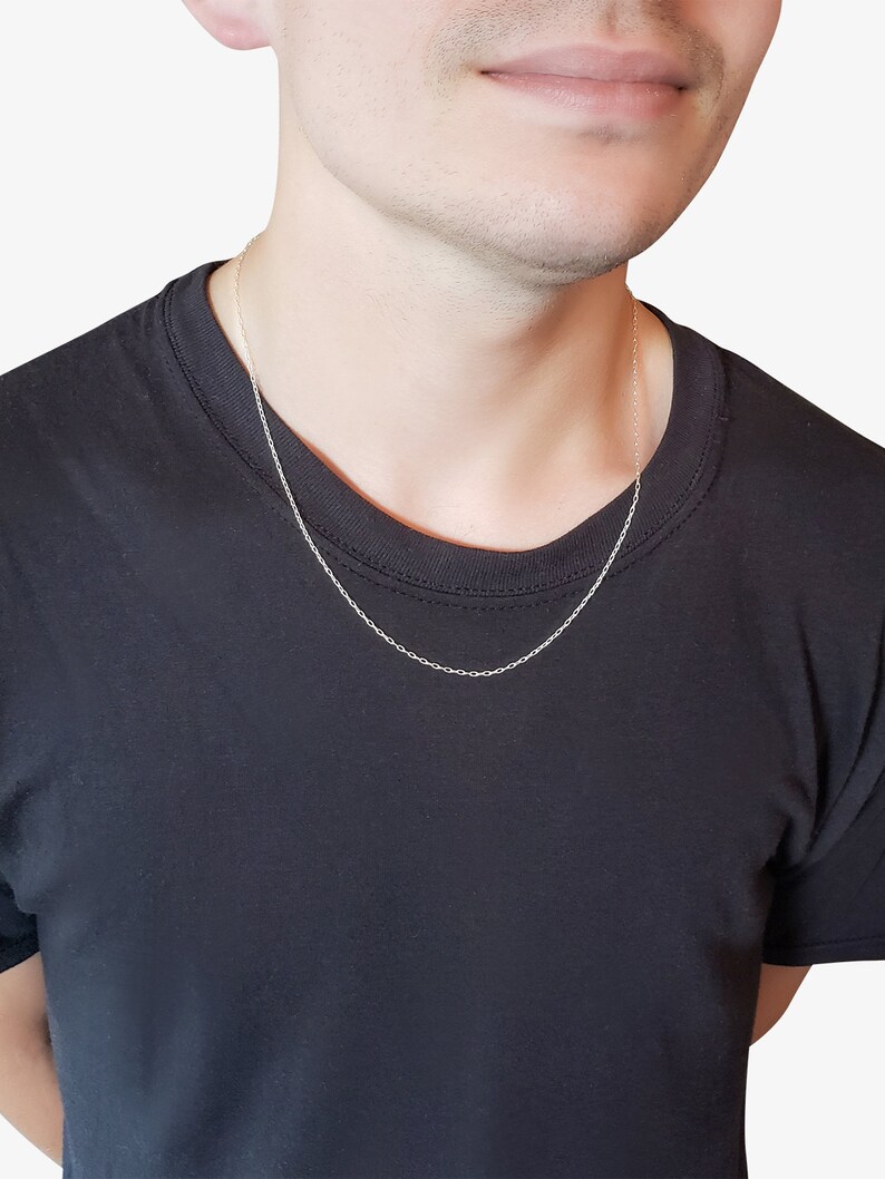 May include: A thin, silver-colored cable chain necklace with small, uniform oval links. The necklace rests against the neck, featuring a simple, delicate design with a polished metallic finish that catches the light, worn over a plain black t-shirt.