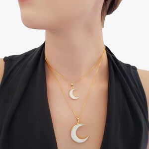 May include: Two gold necklaces with crescent moon pendants. The pendants have a white enamel inlay. The necklaces are layered, with one pendant smaller than the other. The model is wearing a black top.
