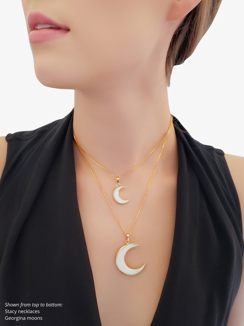 May include: Two gold necklaces with crescent moon pendants. The pendants have a white enamel inlay. The necklaces are layered, with one pendant smaller than the other. The model is wearing a black top.
