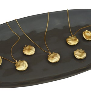 May include: Gold-colored seashell pendant necklaces and charms displayed on a dark gray oval plate. The necklaces have delicate chains, and the pendants are detailed with ribbed textures. The jewelry is arranged against a white background.