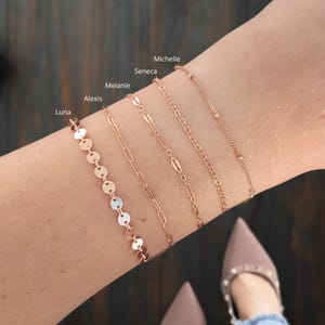 Puede incluir: Cinco pulseras doradas apiladas en la mu&ntilde;eca de una persona. Las pulseras est&aacute;n etiquetadas como "Luna", "Alexis", "Melanie", "Seneca" y "Michelle".