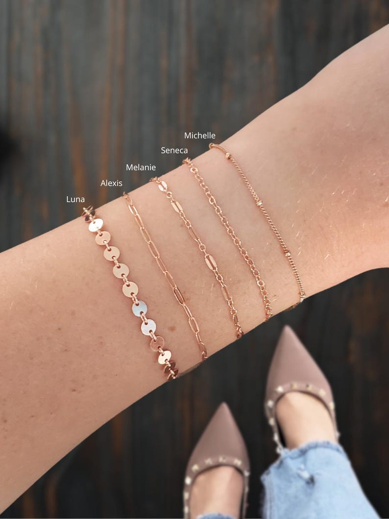 Puede incluir: Cinco pulseras doradas apiladas en la mu&ntilde;eca de una persona. Las pulseras est&aacute;n etiquetadas como "Luna", "Alexis", "Melanie", "Seneca" y "Michelle".