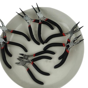 May include: A set of eight black handled pliers with red accents on a white plate. The pliers are used for jewelry making.