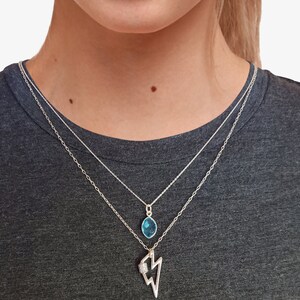 May include: A layered silver necklace set featuring two distinct chains. The shorter chain holds a small, oval-shaped blue gemstone pendant. The longer chain features a silver lightning bolt charm with a small, textured, rectangular accent.