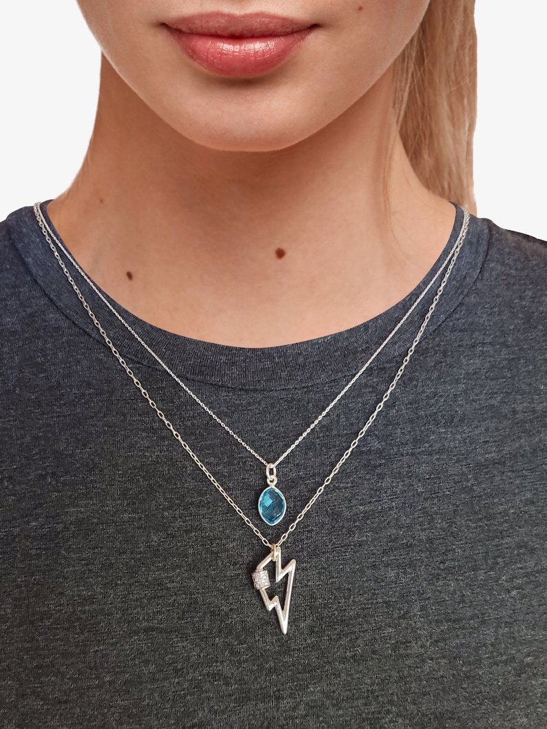 May include: A layered silver necklace set featuring two distinct chains. The shorter chain holds a small, oval-shaped blue gemstone pendant. The longer chain features a silver lightning bolt charm with a small, textured, rectangular accent.