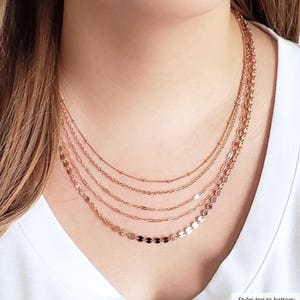 Puede incluir: Un conjunto de collares superpuestos con cinco estilos de cadena diferentes, que incluyen una cadena de cuentas, una cadena de cable, una cadena de lentejuelas, una cadena de clip de papel y una cadena de disco. Las cadenas son todas de color oro rosa.