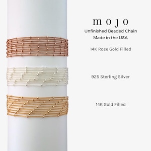 May include: Three beaded chain bracelets displayed on a white cylinder. The top bracelet is rose gold filled, the middle bracelet is sterling silver, and the bottom bracelet is gold filled. The text "mojo" is displayed above the bracelets, followed by "Unfinished Beaded Chain Made in the USA". The text "14K Rose Gold Filled" is displayed below the top bracelet, "925 Sterling Silver" is displayed below the middle bracelet, and "14K Gold Filled" is displayed below the bottom bracelet.