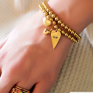 May include: A gold bead bracelet with a heart charm featuring an evil eye design. The bracelet is layered with another gold bead bracelet. A smaller heart charm with a padlock design hangs from the bracelet. The bracelet is worn on a person's wrist.