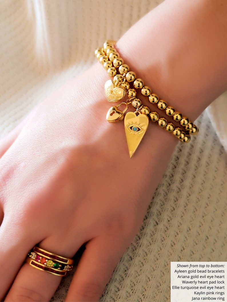 May include: A gold bead bracelet with a heart charm featuring an evil eye design. The bracelet is layered with another gold bead bracelet. A smaller heart charm with a padlock design hangs from the bracelet. The bracelet is worn on a person's wrist.