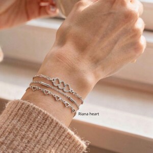 May include: Three silver bracelets with heart-shaped charms and bar accents are stacked on a wrist. The bracelets are delicate and shiny. The text "Riana heart" is visible on the wrist.