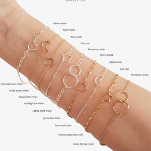 May include: A close-up of a woman's wrist wearing a variety of gold and silver chain bracelets with different charms, including hearts, moons, stars, and circles. The bracelets are labeled with names like "Kenna heart", "Karly moon", "Rory circle", "Cressida heart chain", "Linda dainty chain", "Kaylani link chain", "Ashleigh bar chain", "Sasha circle chain", "Jackie bar chain", "Starr oval chain", "Celeste paperclip chain", and "Desie flat bar chain".