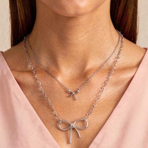 May include: Two silver necklaces with bow pendants. The top necklace has a small bow, while the bottom necklace features a larger bow. The top necklace is a cable chain, and the bottom necklace is a heart chain. The image also includes the text: "Styles top to bottom: Eloise cable chain, Leona small bow, Christie heart chain, Indie large bow."