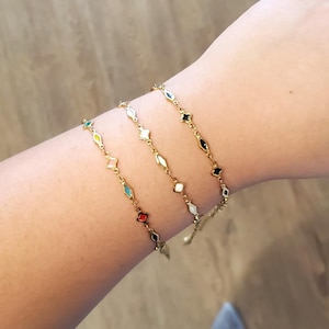 K&ouml;nnte beinhalten: Drei goldfarbene Armb&auml;nder mit farbenfrohen geometrischen Anh&auml;ngern werden gezeigt. Jedes Armband hat eine zarte Kette und eine Reihe von rauten- und sternf&ouml;rmigen Anh&auml;ngern in verschiedenen Farben, darunter Rot, Schwarz, Wei&szlig; und T&uuml;rkis.