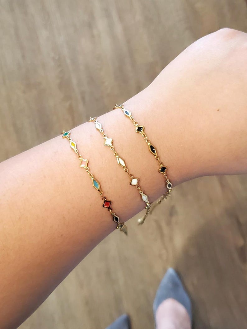 K&ouml;nnte beinhalten: Drei goldfarbene Armb&auml;nder mit farbenfrohen geometrischen Anh&auml;ngern werden gezeigt. Jedes Armband hat eine zarte Kette und eine Reihe von rauten- und sternf&ouml;rmigen Anh&auml;ngern in verschiedenen Farben, darunter Rot, Schwarz, Wei&szlig; und T&uuml;rkis.
